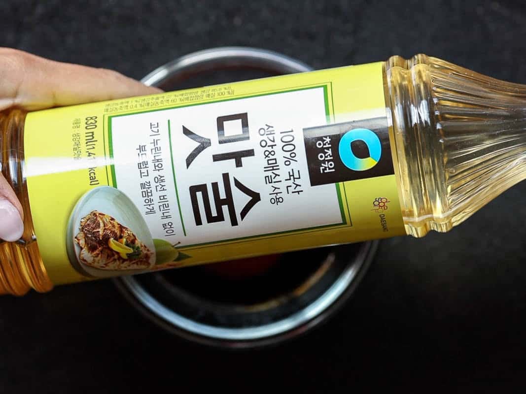 A hand holding a Korean mirin bottle over a stainless steel bowl filled with dark liquid. The bottle has a yellow label with text and an image of a dish. The background is a dark surface.